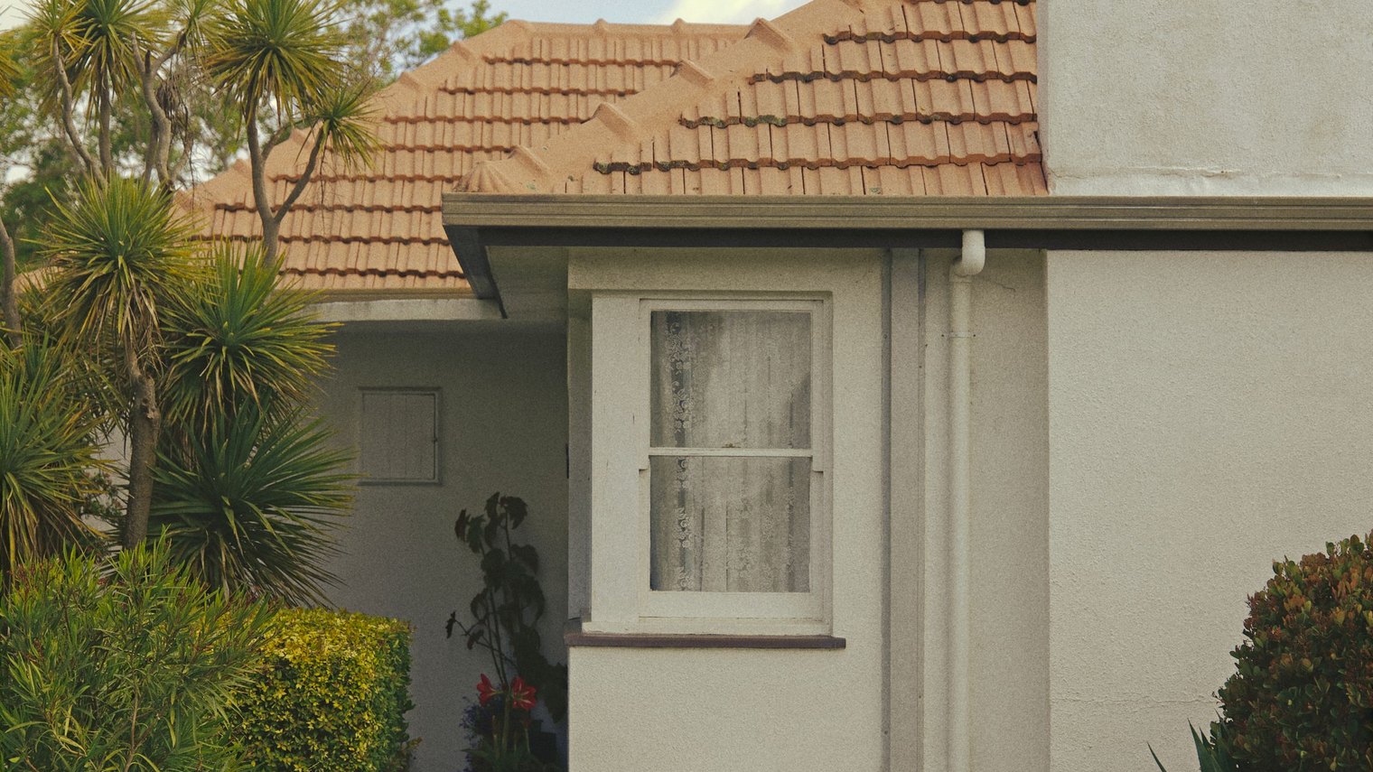 A modest rendered Australian house with a terracotta tile roof, an air conditioning unit and a cracked front fence.
