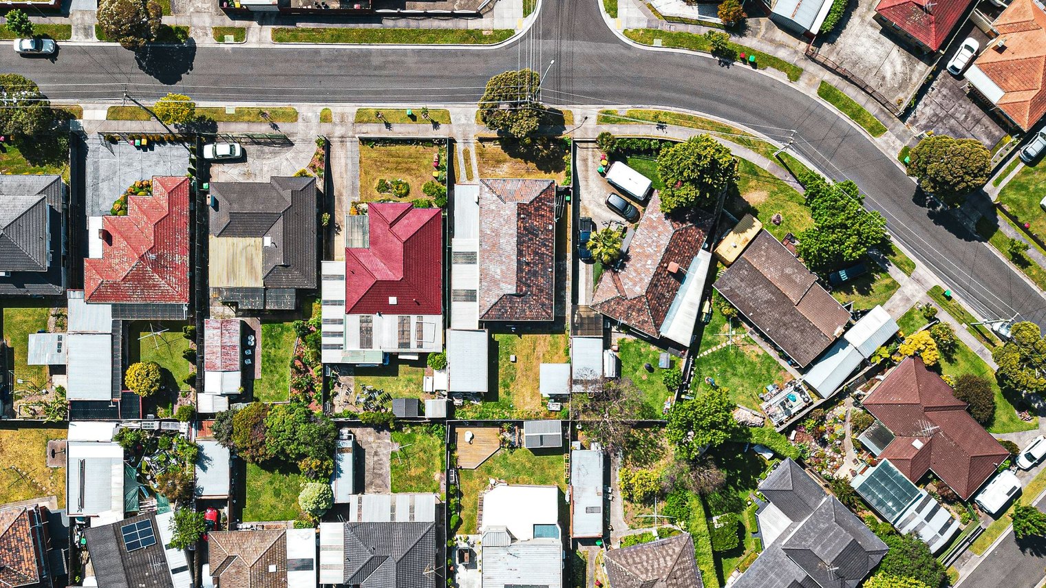 Aerial view looking straight down over an established Australian suburb, a patchwork of tile and metal roofs, backyards, solar panels and quiet streets.