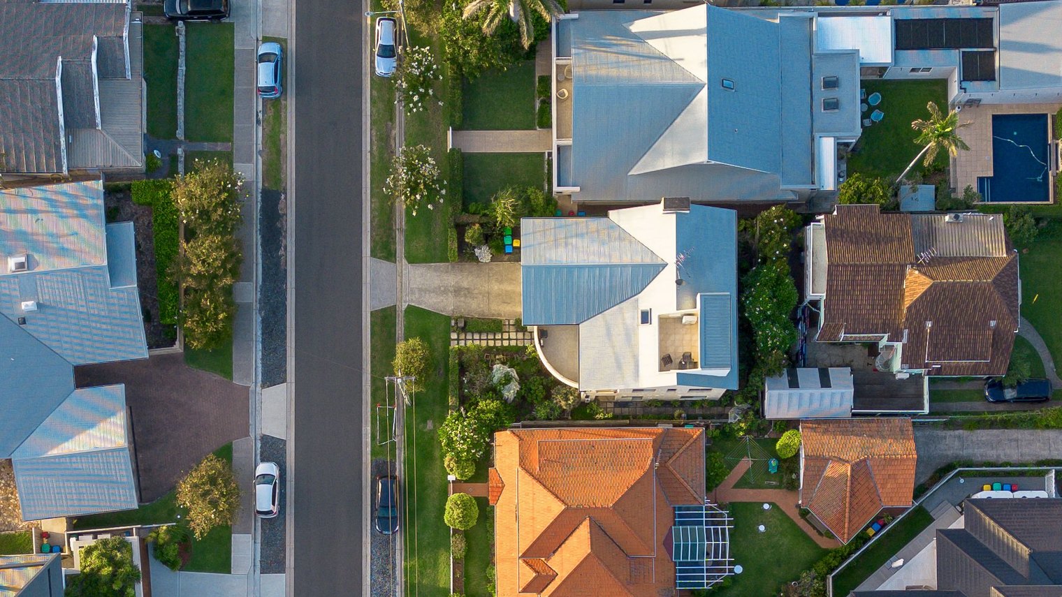 Aerial view looking down a quiet Australian residential street of tile roofed homes with pools and solar panels.